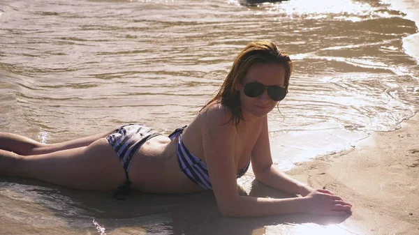 Blondes Mädchen im Badeanzug und schwarzer Sonnenbrille an einem weißen Sandstrand am Meer im Urlaub. schönes Model mit sexy Körper schwimmt im blauen Meerwasser — Stockfoto