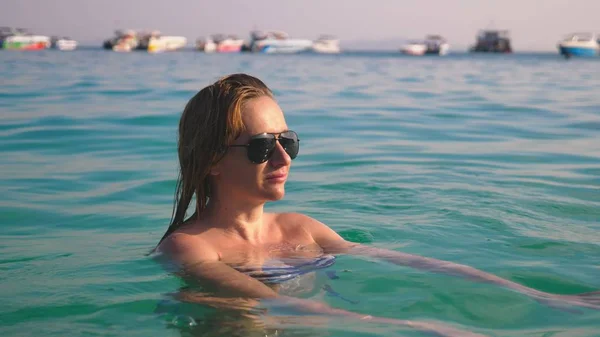 Gadis pirang dalam pakaian renang dan kacamata hitam. Model indah dengan tubuh seksi berenang di air laut biru — Stok Foto