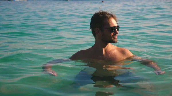 Retrato de um jovem bonito com barba em óculos de sol relaxado em água cristalina do mar — Fotografia de Stock