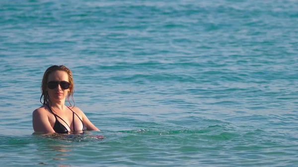 A menina a loira em um maiô preto e óculos pretos. Modelo bonito com corpo sexy está se divertindo na água do mar cristalina . — Fotografia de Stock
