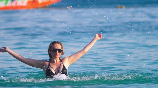 La fille la blonde en maillot de bain noir et des lunettes noires. Beau modèle avec corps sexy s'amuse dans de l'eau de mer cristalline . — Photo