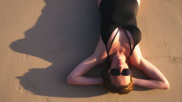 Blond flicka i en svart baddräkt och svarta solglasögon på en vit sandstrand. Vacker modell med sexig kropp Solar vid havet — Stockfoto