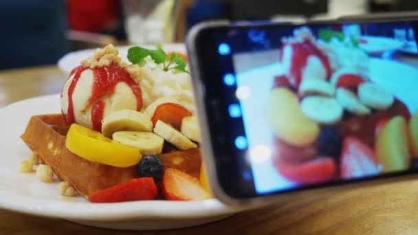 Close-up. As mãos femininas tiram uma foto de uma sobremesa de waffles vienenses, sorvete e frutas frescas em um smartphone em um restaurante. . — Vídeo de Stock