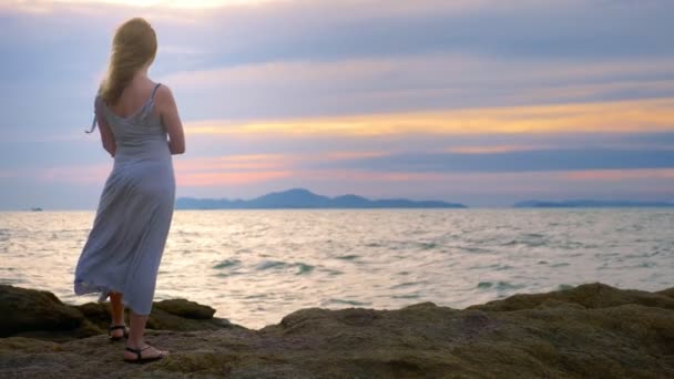 Mujer joven caminando sobre una roca y disfrutando de la vista de la puesta del sol, salpicando las olas — Vídeos de Stock
