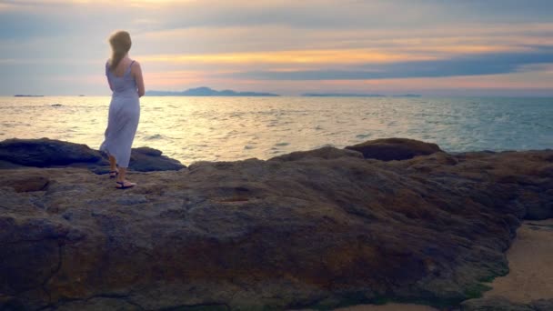 Mujer joven caminando sobre una roca y disfrutando de la vista de la puesta del sol, salpicando las olas — Vídeos de Stock