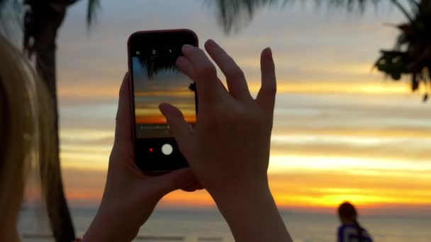 En kvinna med en mobiltelefon gör ett foto av en tropisk strand i solnedgången. — Stockvideo