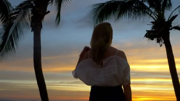 Menina loira fica ao lado da palmeira na praia ao pôr do sol — Vídeo de Stock