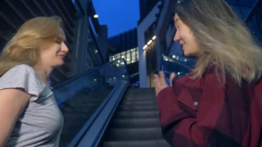 iki kız, Asya ve beyaz, modern kent sokak akşamları sohbet edin. yürüyen merdiven üzerinde duran.