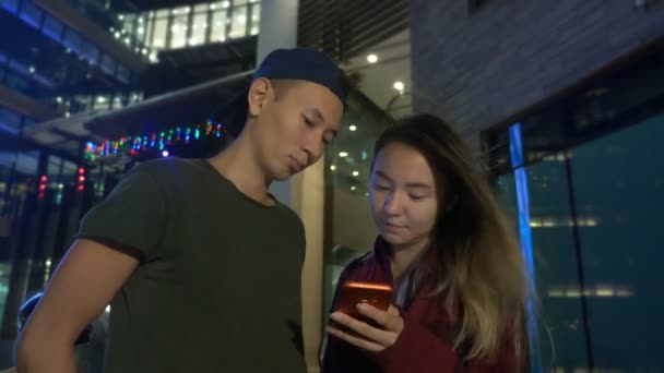 快乐的年轻的亚洲夫妇的男朋友和女朋友。晚上站在城市街道上使用智能手机. — 图库视频影像