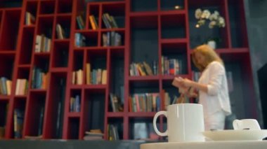 Kütüphanede bir masada sıcak bir içecek ile bir kupa üzerinde duruluyor. Bulanıklık, okumak için bir kitap seçerek, bir kitaplık önünde duran genç kadın,