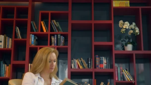 Mujer joven leyendo un libro en la biblioteca en el fondo de las estanterías — Vídeos de Stock