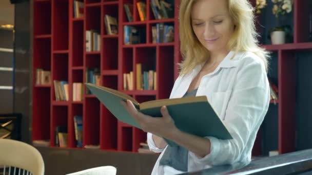 Молода жінка читає книгу в бібліотеці на фоні книжкових полиць — стокове відео