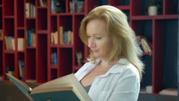 Mujer joven leyendo un libro en la biblioteca en el fondo de las estanterías — Vídeos de Stock