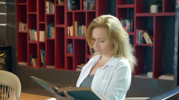 Mujer joven leyendo un libro en la biblioteca en el fondo de las estanterías — Vídeos de Stock