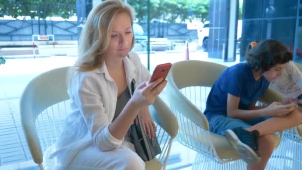 Een drukke vrouw maakt gebruik van de telefoon in de lobby van het hotel, haar kinderen naast haar zitten en ook gebruik maken van de telefoon — Stockvideo