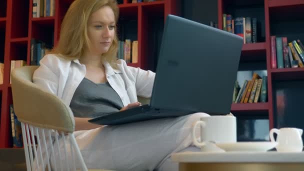젊은 여자는 웃 고. 현대 도서관에 앉아있는 동안 노트북을 사용 하 여. — 비디오