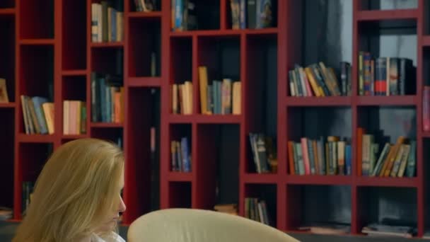 Genç kadın gülümsüyor. modern kitaplığa otururken bir dizüstü bilgisayar kullanarak. — Stok video