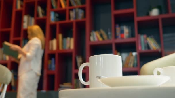 Focusing on a mug with a hot drink, on a table in the library. Blur, Young woman, choosing a book to read, standing in front of a bookcase. — Stock Video