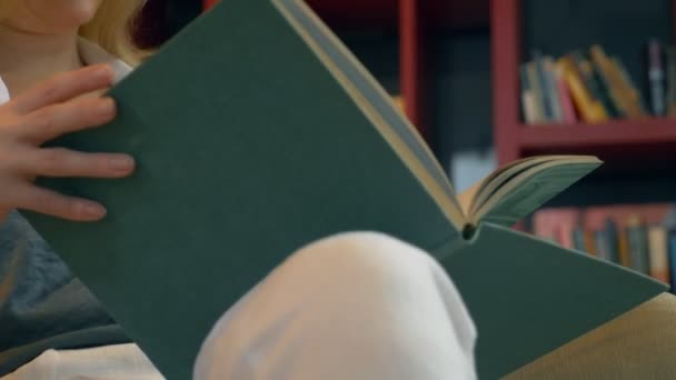 Mujer joven leyendo un libro en la biblioteca en el fondo de las estanterías — Vídeos de Stock