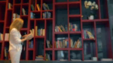 Kütüphanede bir masada sıcak bir içecek ile bir kupa üzerinde duruluyor. Bulanıklık, okumak için bir kitap seçerek, bir kitaplık önünde duran genç kadın,.