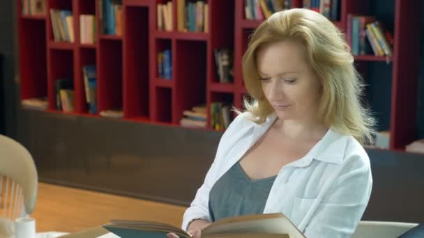 Mujer joven leyendo un libro en la biblioteca en el fondo de las estanterías — Vídeos de Stock