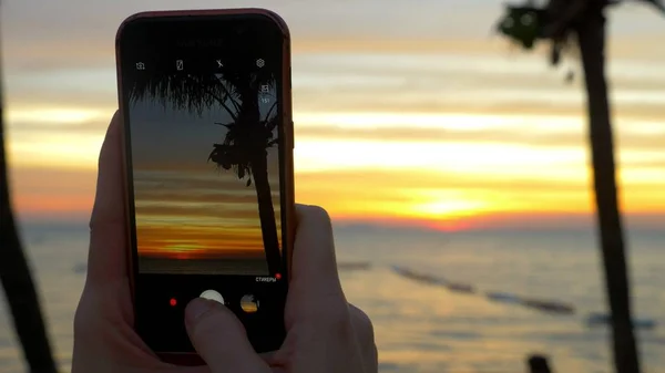 Uma mulher com um telefone celular faz uma foto de uma praia tropical ao pôr do sol . — Fotografia de Stock