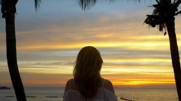 Menina loira fica ao lado da palmeira na praia ao pôr do sol — Fotografia de Stock