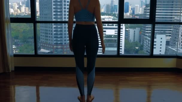 Mujer joven haciendo yoga en una habitación cerca de una gran ventana con vistas a los rascacielos . — Vídeos de Stock
