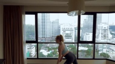 Genç kadın gökdelenler bakan büyük bir pencere yakınında bir odada yoga yaparken.