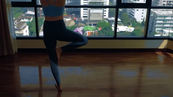 Mujer joven haciendo yoga en una habitación cerca de una gran ventana con vistas a los rascacielos . — Vídeos de Stock