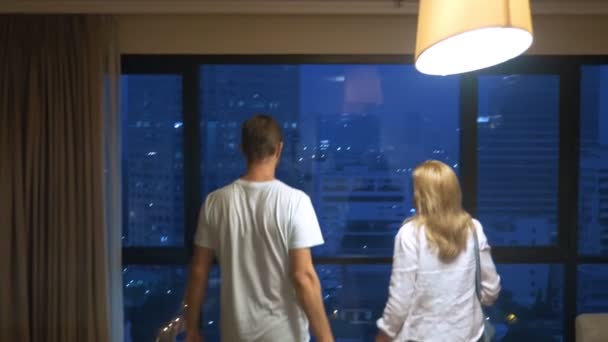 Attraktives Paar, Frau und Mann mit einem Koffer vor dem Hintergrund von Wolkenkratzern in einem Panoramafenster am Abend — Stockvideo