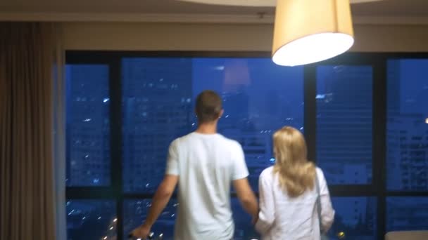 Pareja atractiva, mujer y hombre con una maleta en el fondo de rascacielos en una ventana panorámica por la noche — Vídeo de stock