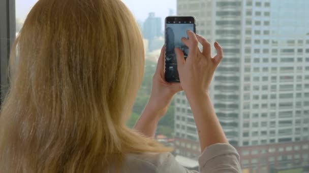 Элегантная блондинка делает фото по телефону. женщина фотографирует вид из окна небоскреба — стоковое видео