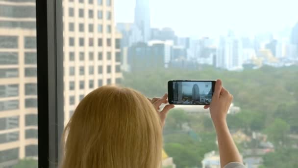 Mujer rubia elegante haciendo una foto en el teléfono. mujer fotografía la vista desde la ventana de los rascacielos — Vídeos de Stock