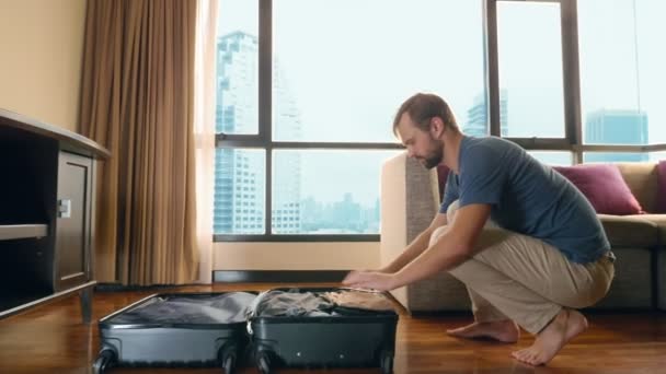 Красивый мужчина упаковывает чемодан в комнате с панорамным окном с видом на небоскребы — стоковое видео
