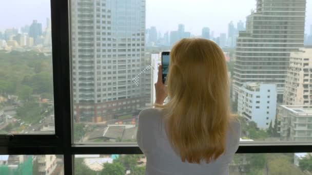 Elegáns szőke nő, ami egy fotót a telefon. nő a kilátás az ablakból a felhőkarcolók fényképek — Stock videók