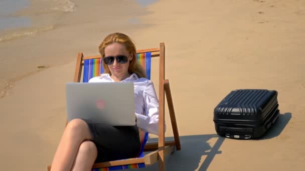 Empresária trabalhando em um laptop enquanto sentado em uma espreguiçadeira junto ao mar em uma praia de areia branca. conceito freelance ou de workaholism . — Vídeo de Stock