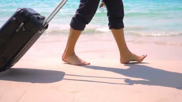Ein junger Mann in Hose und klassischem Hemd mit Koffer spaziert vor der Kulisse des türkisfarbenen Meeres am Strand entlang. freiberufliches Konzept, Ruhe — Stockvideo