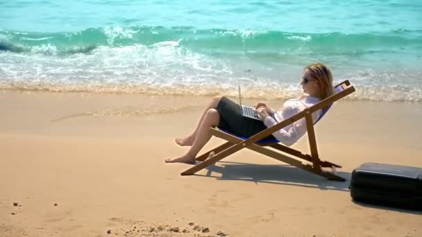Zakenvrouw werking voort te laptop zittend in een ligstoel aan zee op een wit zandstrand. freelance of workaholism concept. — Stockvideo