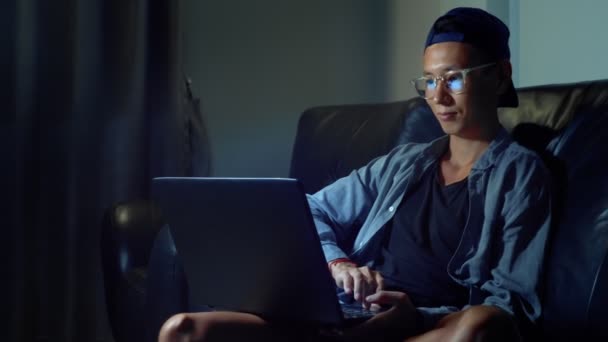 Jonge knappe Aziatische man in glazen met reflecties met behulp van zijn laptop, zitten in de avond in de kamer. In het donker — Stockvideo
