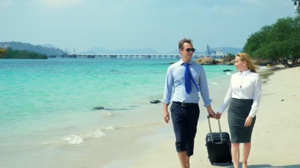 Бизнесмен и деловая женщина с чемоданом прогуливаются по пляжу с белым песком на острове — стоковое видео