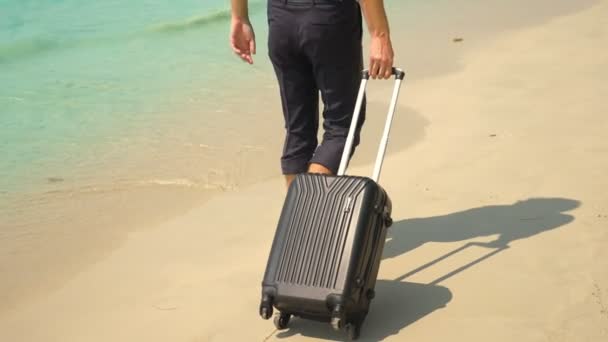 Ein junger Mann in Hose und klassischem Hemd mit Koffer spaziert vor der Kulisse des türkisfarbenen Meeres am Strand entlang. freiberufliches Konzept, Ruhe — Stockvideo