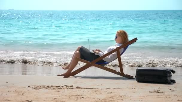 Femme d'affaires travaillant sur un ordinateur portable assis dans une chaise longue au bord de la mer sur une plage de sable blanc. concept freelance ou workaholism . — Video