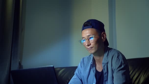Joven hombre asiático guapo en gafas con reflejos usando su computadora portátil, sentado en la noche en la habitación. En la oscuridad — Vídeos de Stock