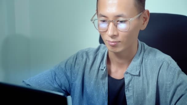 Όμορφος Ασίας νεαρός χρησιμοποιώντας φορητό υπολογιστή του, κάθεται στο γραφείο σε γραφείο — Αρχείο Βίντεο