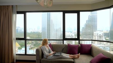 zarif bir kadın gökdelenler bakan ve onu laptop kullanarak panoramik cam kenarında bir kanepede yatıyor