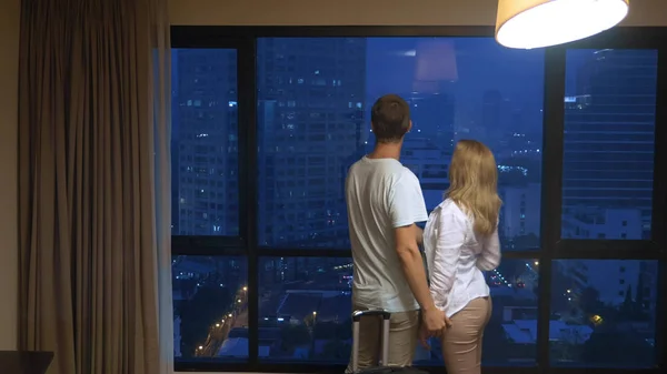 Привлекательная пара, женщина и мужчина с чемоданом на фоне небоскребов в панорамном окне вечером — стоковое фото