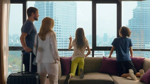 Familia feliz, mujer, hombre y dos niños con una maleta en el fondo de rascacielos en una ventana panorámica — Foto de Stock