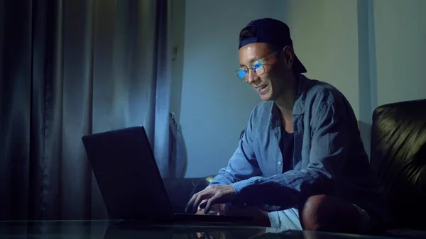 Jeune homme asiatique beau dans des lunettes avec des reflets à l'aide de son ordinateur portable, assis le soir dans la chambre. Dans le noir — Photo