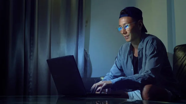 Jeune homme asiatique beau dans des lunettes avec des reflets à l'aide de son ordinateur portable, assis le soir dans la chambre. Dans le noir — Photo